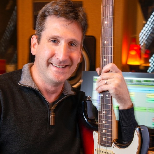 Scott Leader holding a guitar in front of a mixing board and computer